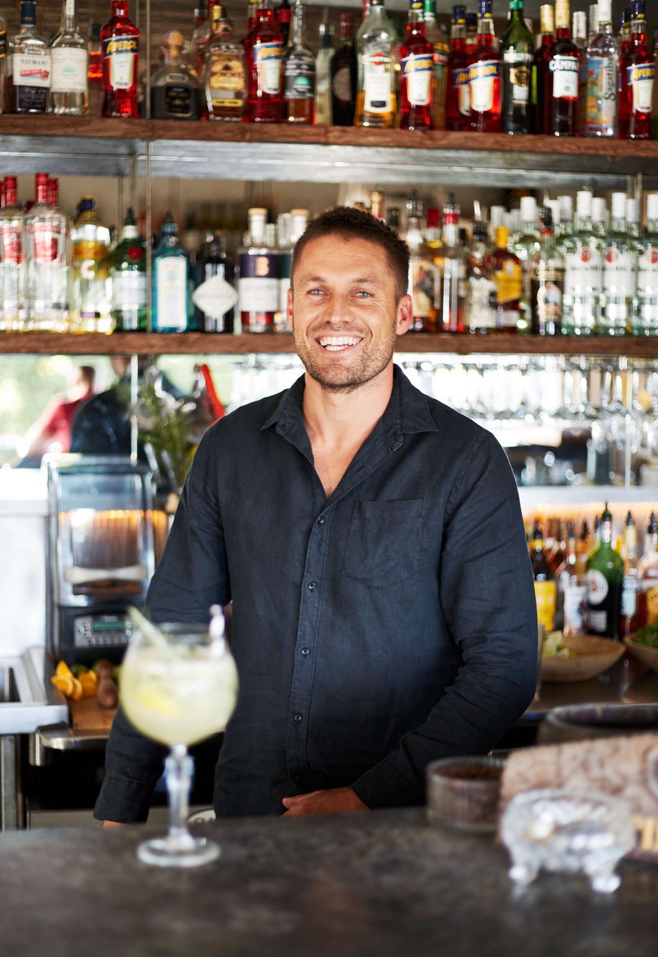 Bartender Jesse Williams smiles behind the bar at Paradiso, with shelves of spirits behind him.