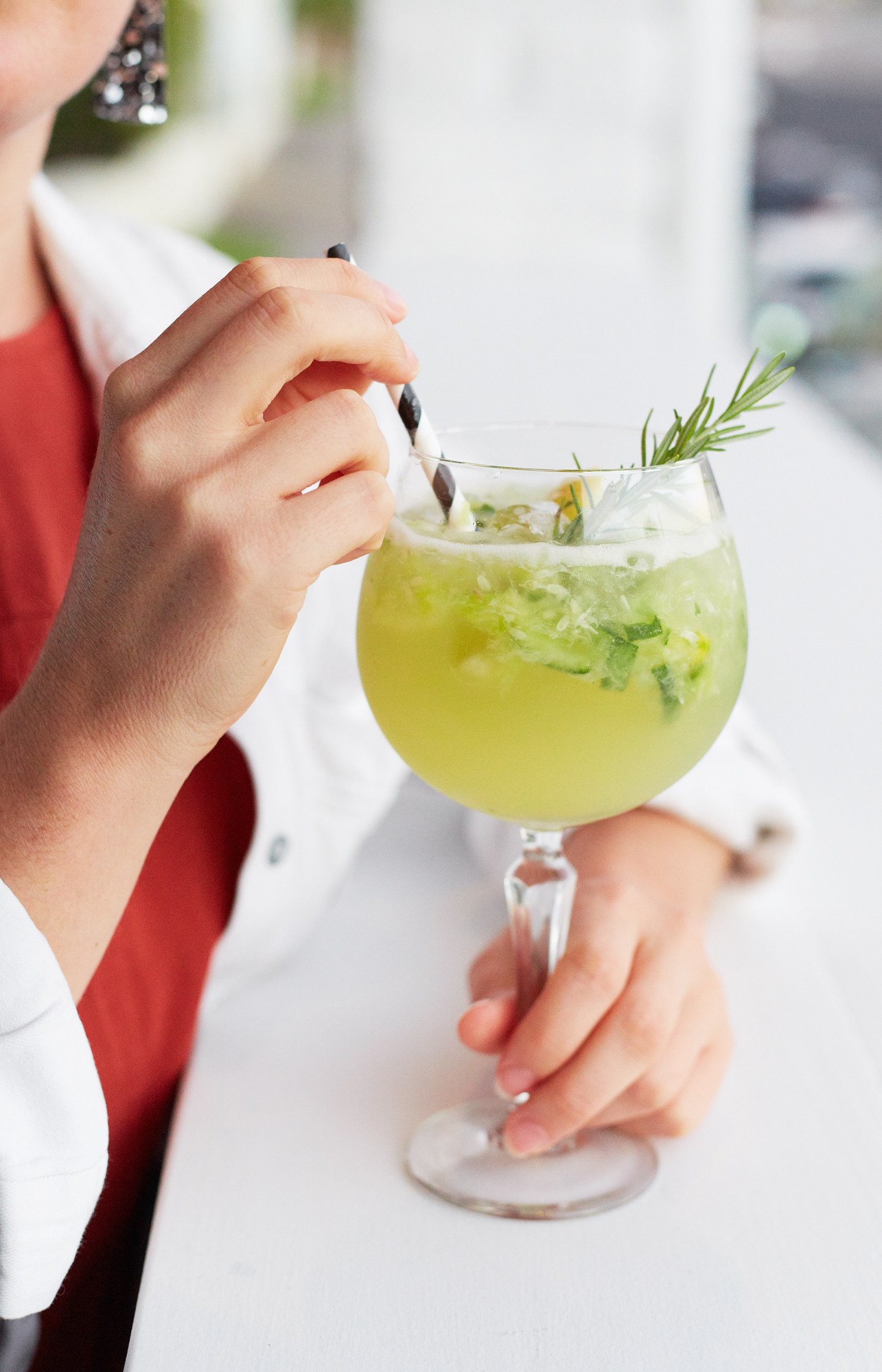 Woman in red and white holding a green cocktail with rosemary garnish and black olives at Paradiso bar.