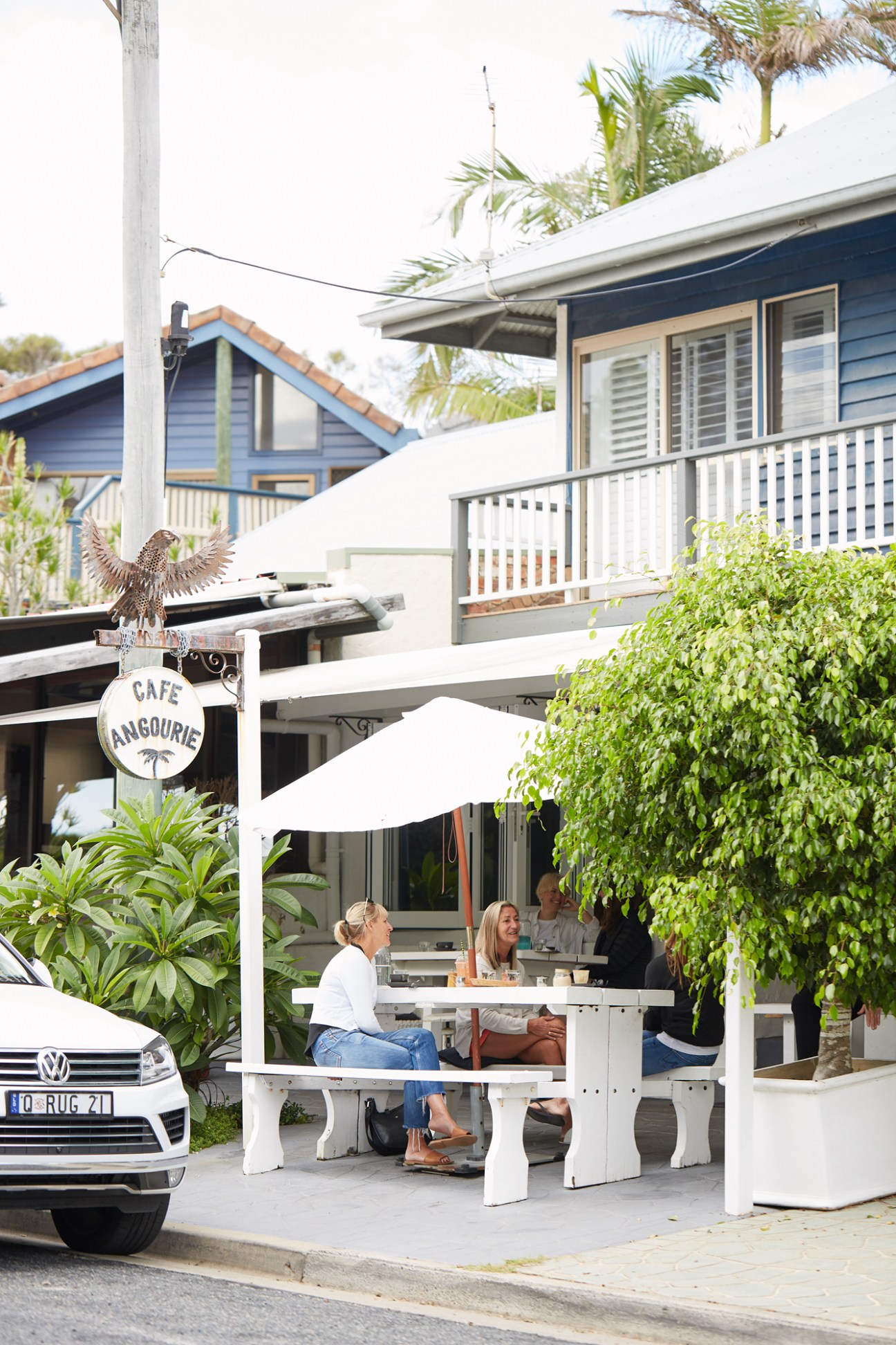 Café Angourie storefront with white umbrellas, diners seated at tables, palm trees, and residential buildings in background.