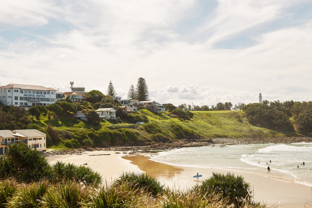 The Pacific Hotel overlooks Main Beach in Yamba with surfers in the water and green hillside landscape.