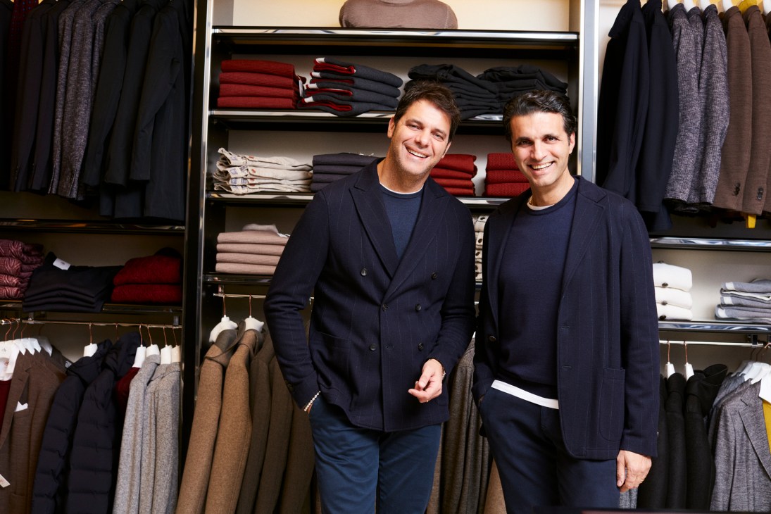 Two men in matching navy blazers and shirts stand smiling in front of shelves displaying folded clothing in various colors.