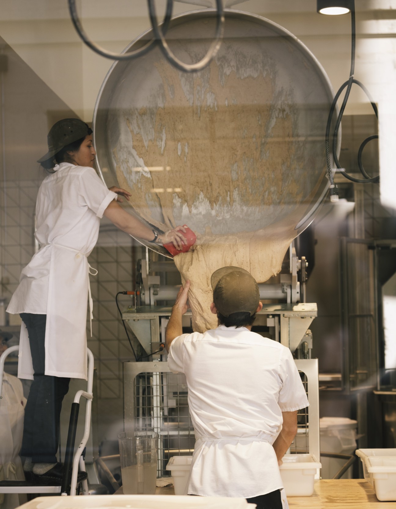 Two bakers in white coats pour dough from a large transparent mixing bowl at The Manufactory bakery in Los Angeles.