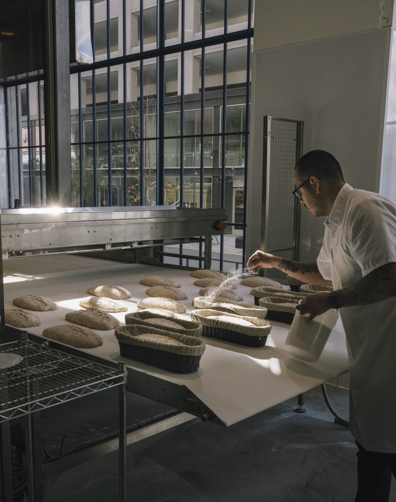 Baker in white shirt dusting flour onto shaped ciabatta loaves at The Manufactory bakery in Los Angeles.