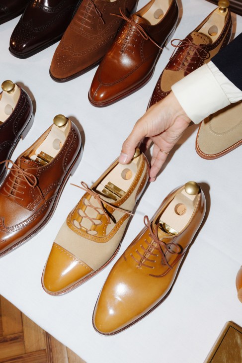A hand holding a wooden shoe tree inside a polished tan and cream oxford shoe surrounded by other dress shoes.