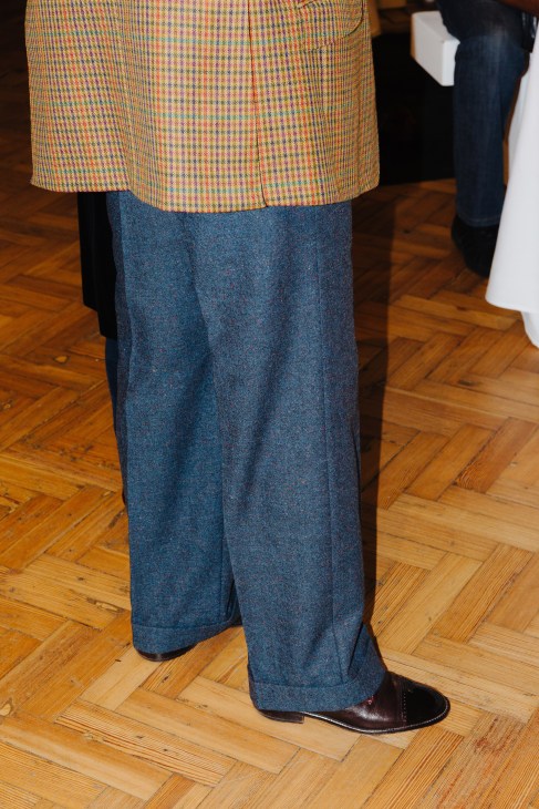 Person wearing a multicolored houndstooth jacket and blue denim trousers with brown leather shoes on a wooden floor.