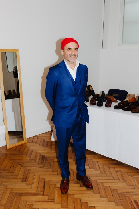Man in navy suit and red cap stands in room with shoes displayed, flexing bicep.