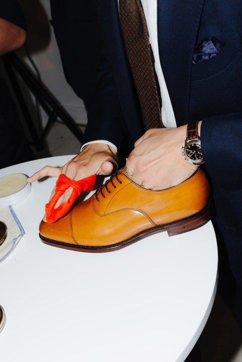Person in navy suit polishing a tan leather oxford shoe with a red cloth while wearing a watch.