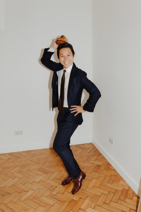 Yuya Hasegawa in a navy suit holds a shoe above his head while posing with a smile.