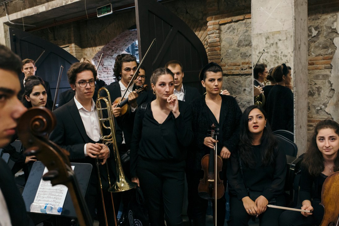 Young musicians in formal black attire gather backstage, holding their instruments and listening intently during a pre-performance briefing.