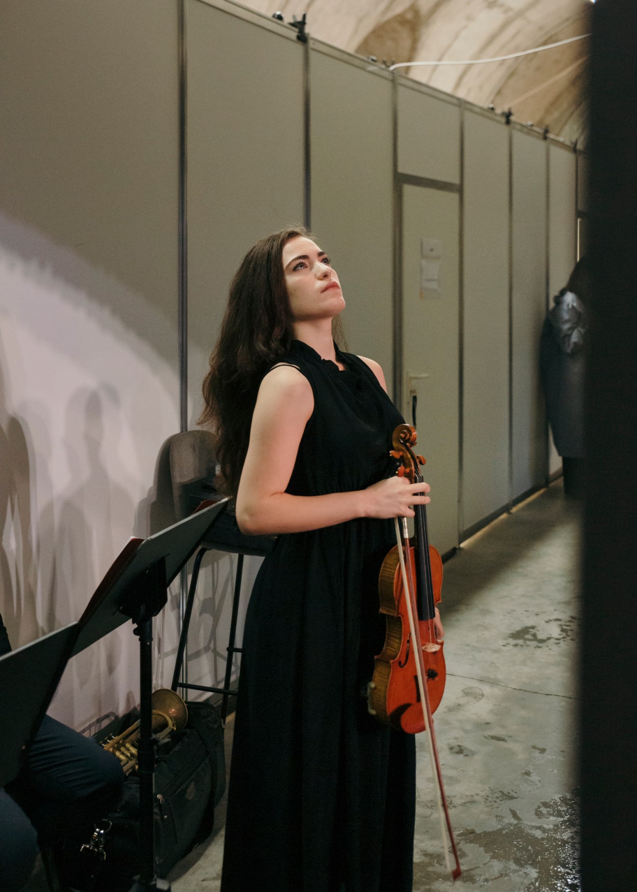 Young female violinist in black dress holding violin, looking upward backstage before performance.