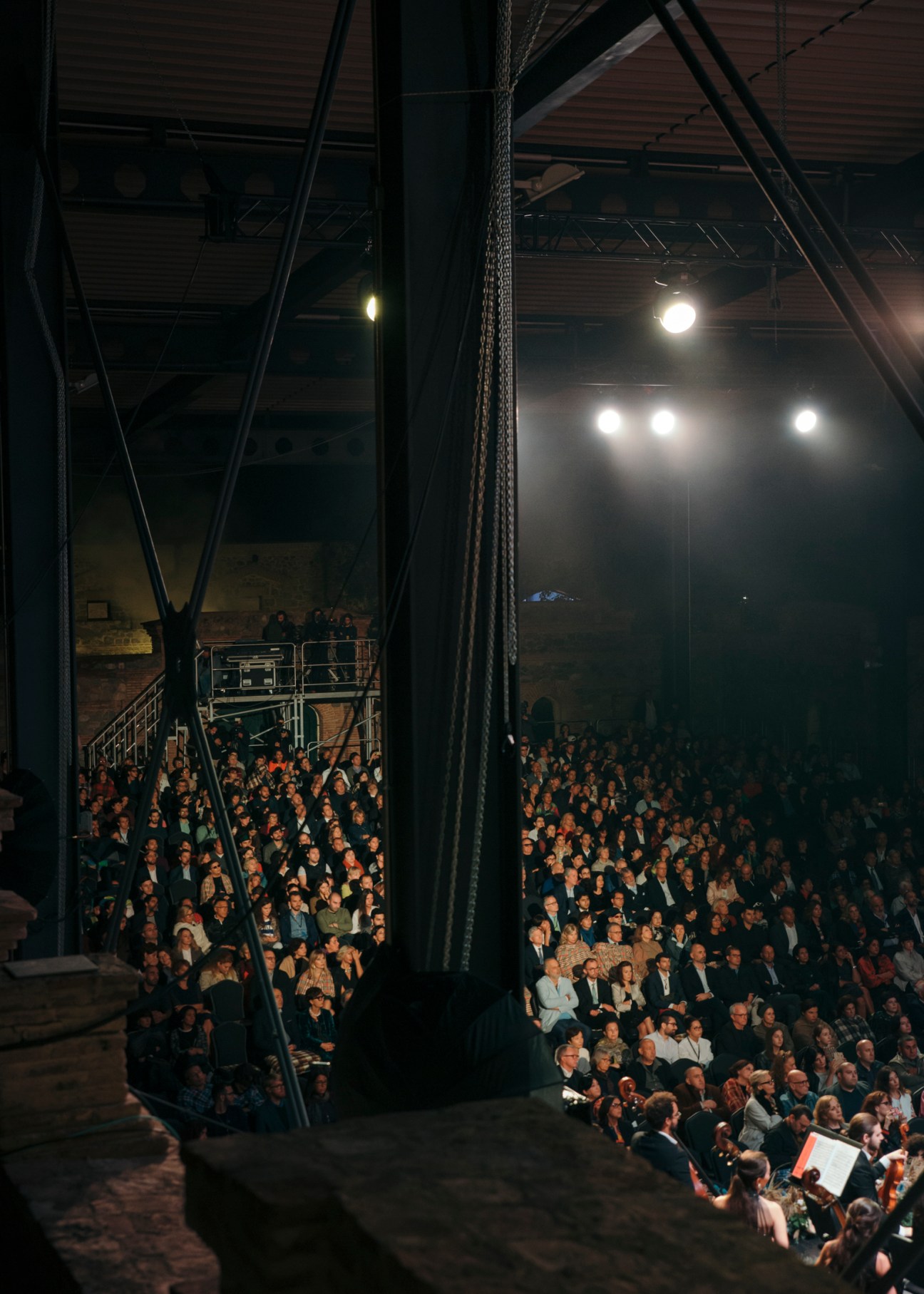 A large audience seated in an industrial concert hall with steel beams and spotlights overhead.