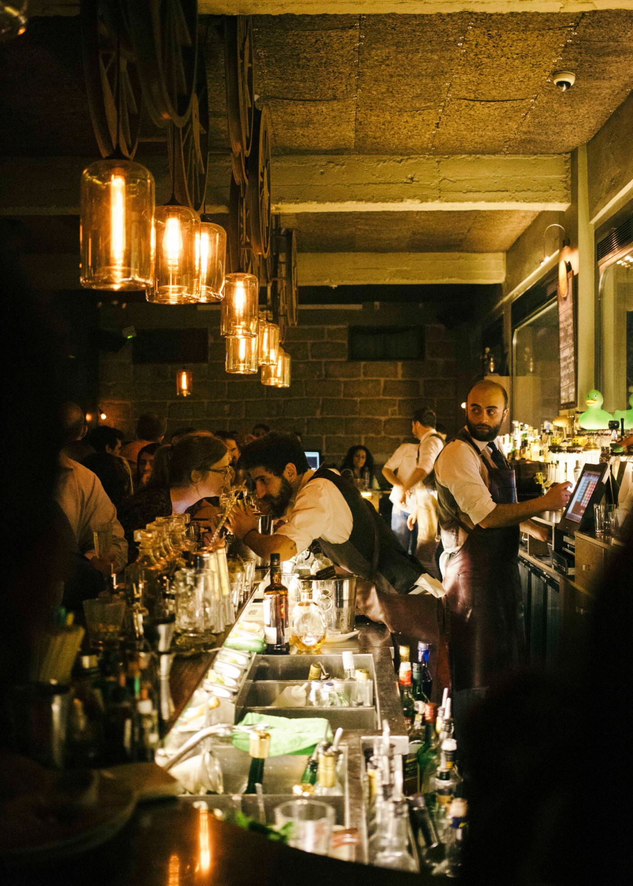 Crowded bar scene with bartender in vest serving drinks under hanging golden lights at night in Beirut.