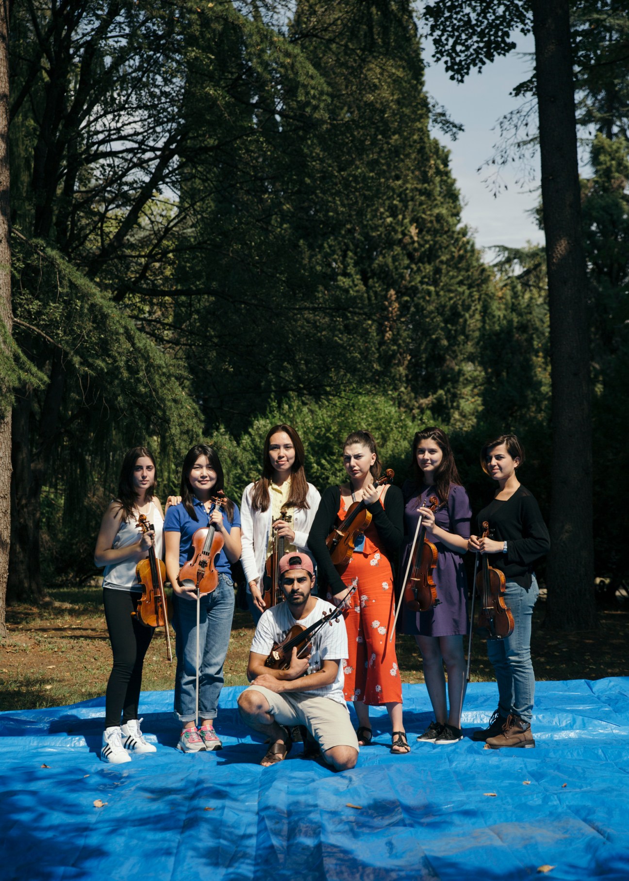 Young musicians holding violins stand together outdoors in a forested setting on a blue platform.