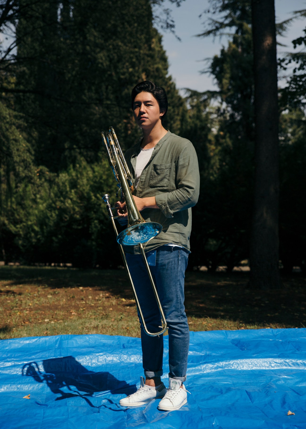 Kazakh bass trombonist Doskhan Zhexembay holds his instrument while standing on a blue tarp in a forest clearing.