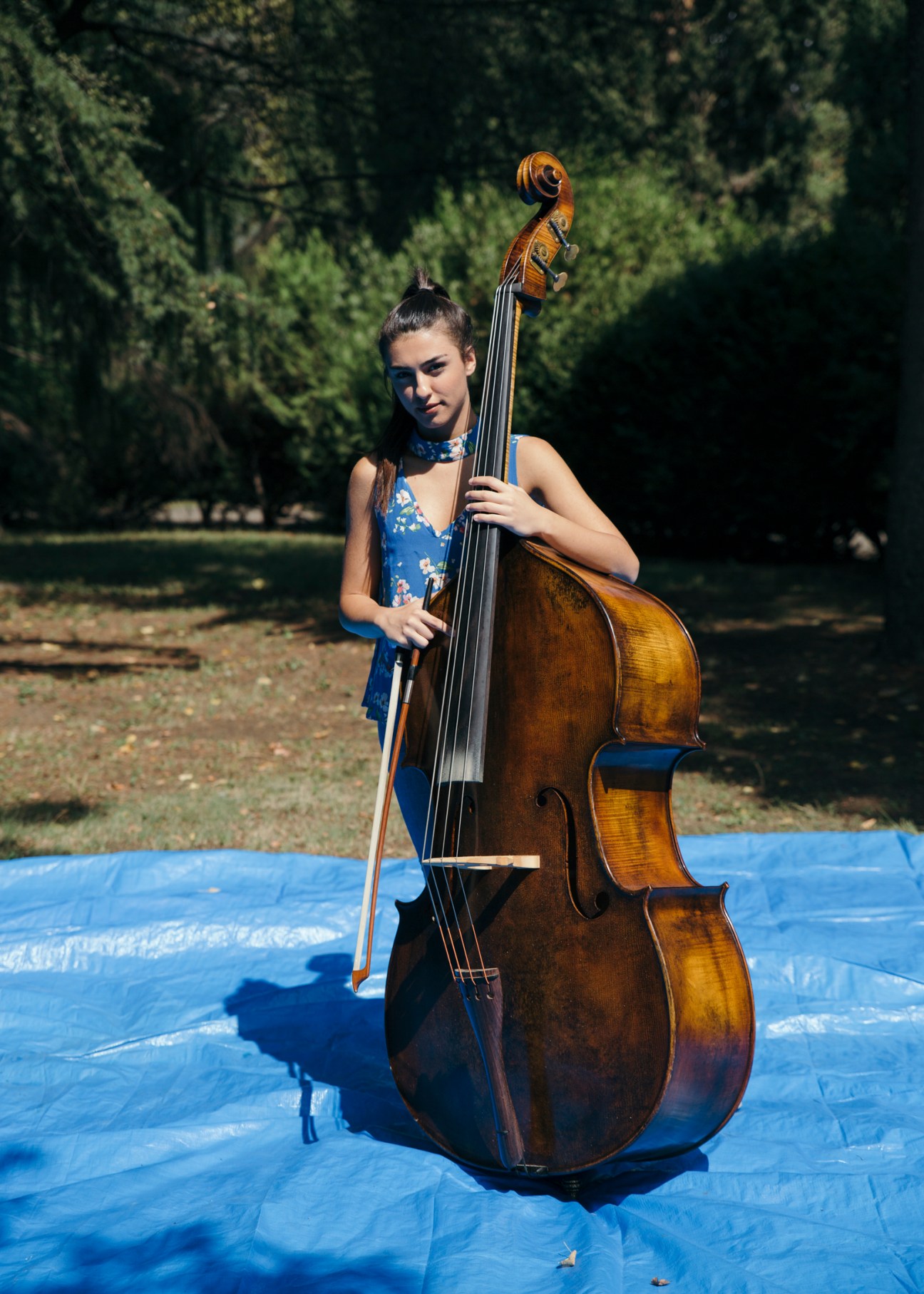 Young female cellist in blue dress playing outdoors on blue mat with forested background.
