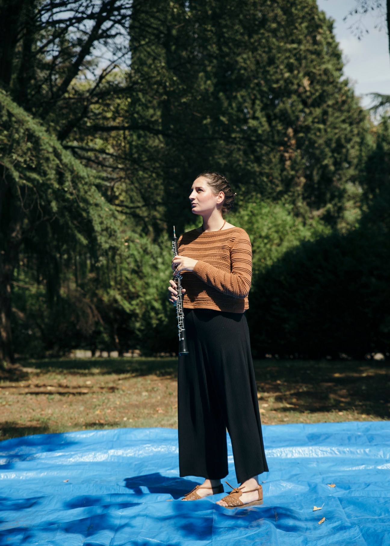 Georgian oboist Ekaterine Tsenderadze holds her instrument while standing on blue tarp surrounded by tall cypress trees.