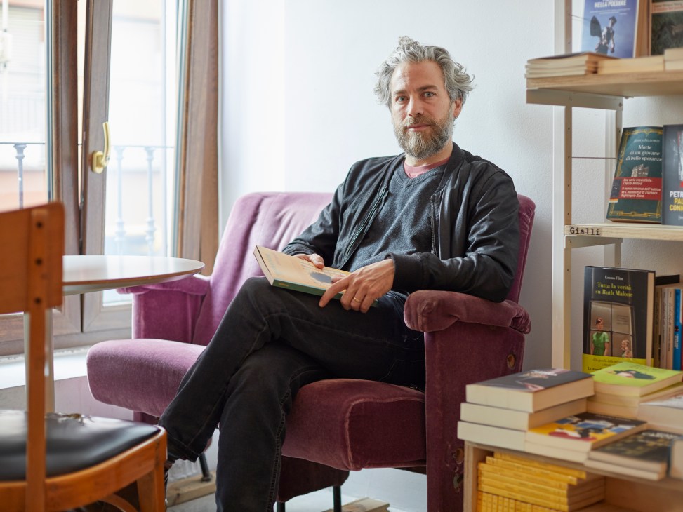 Pietro Biancardi sitting in a burgundy armchair holding a book, surrounded by bookshelves in a bright office