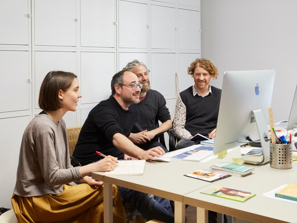 Four people collaborating around a desk with books, papers and an iMac computer in a bright office