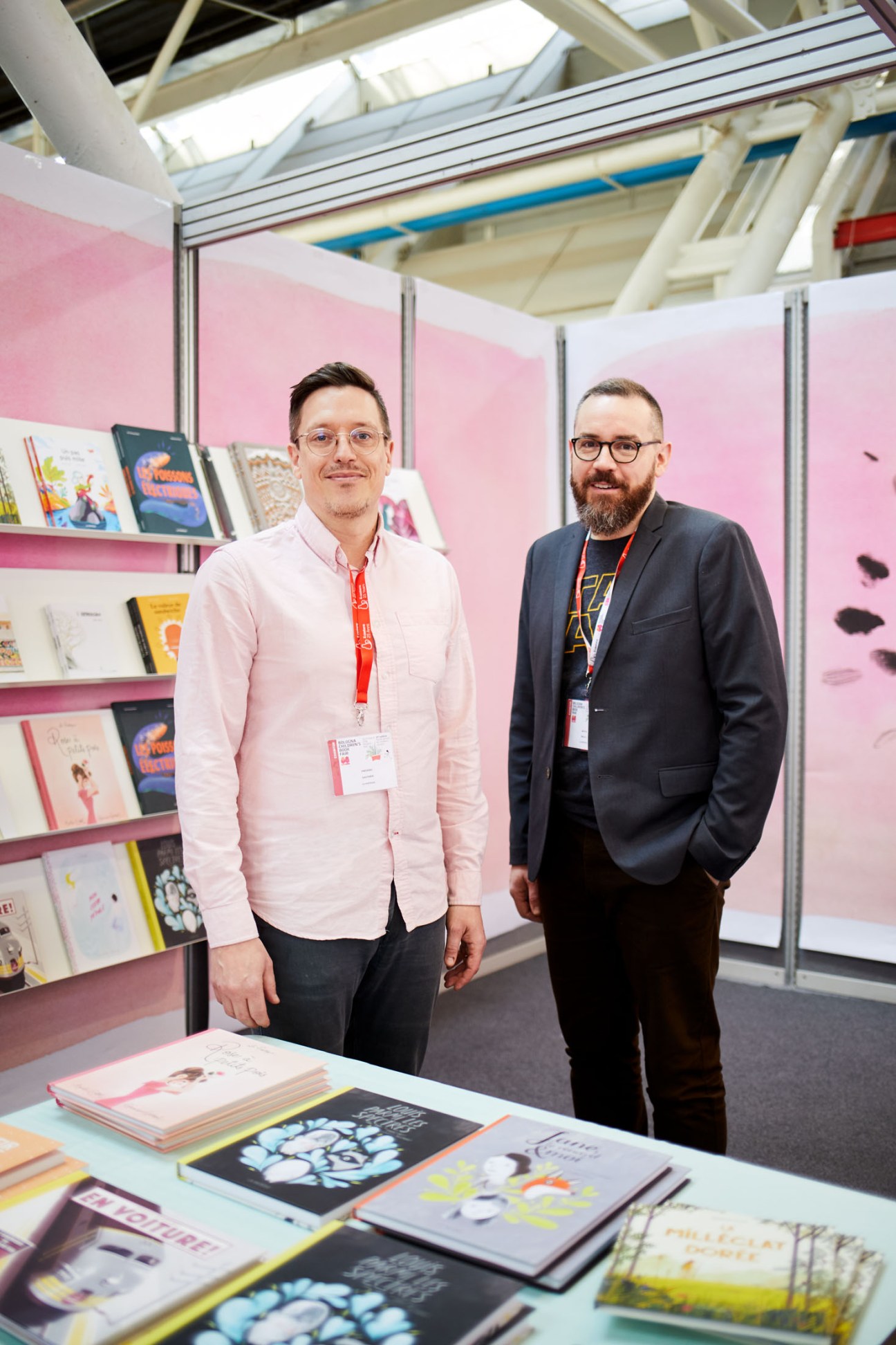 Two men standing in a pink-walled exhibition booth with bookshelves displaying children's books at Bologna's Children's Book Fair.