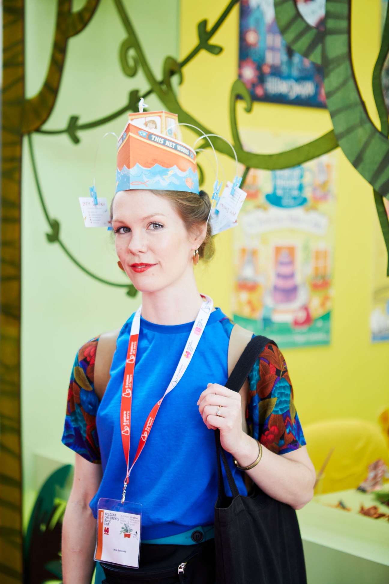 Woman wearing a children's book-themed paper boat hat and lanyard at Bologna's Children's Book Fair.