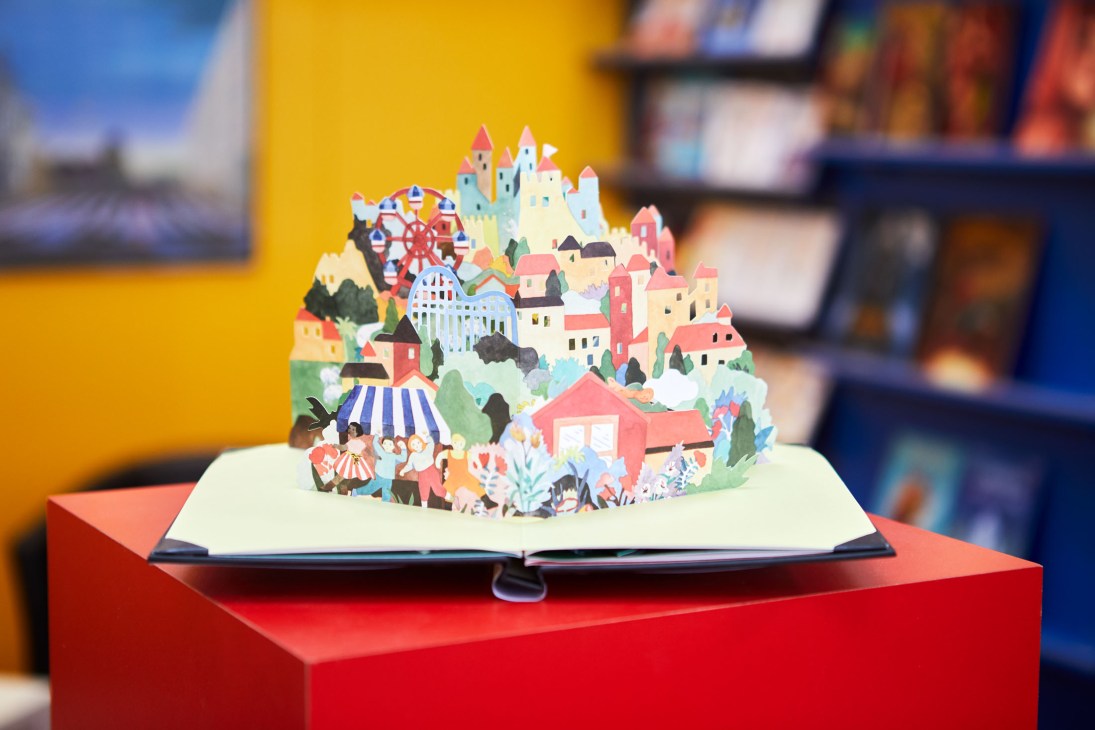 An open pop-up children's book displaying a colorful three-dimensional village with houses, castle, and figures on a red display pedestal.