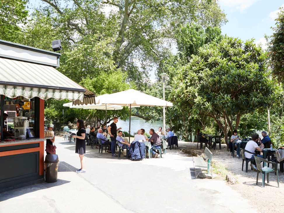 Outdoor riverside kiosk and café with white umbrellas where people dine and socialize along Turin's riverbank.