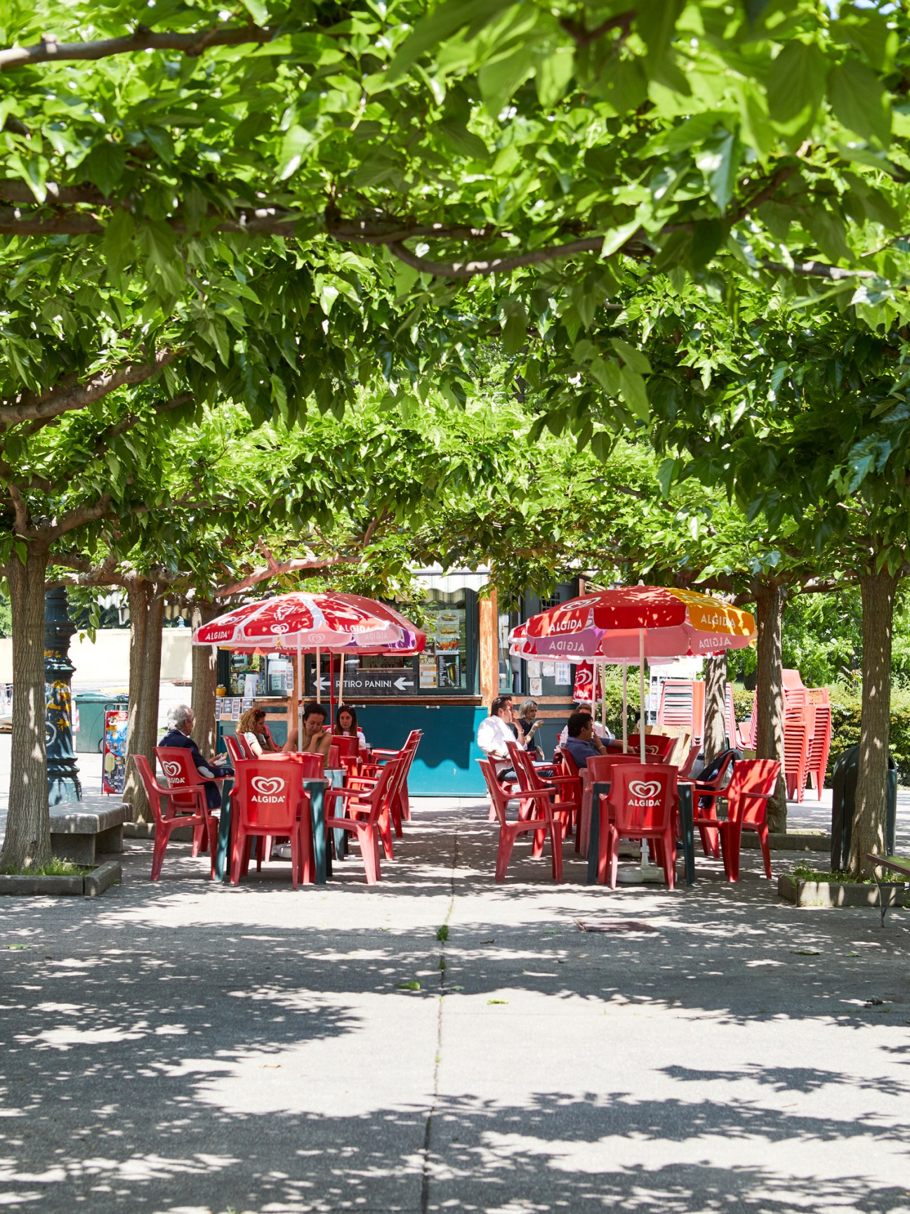 Red umbrellas and chairs at a typical kiosk in Parco del Valentino, Turin, shaded by green trees.