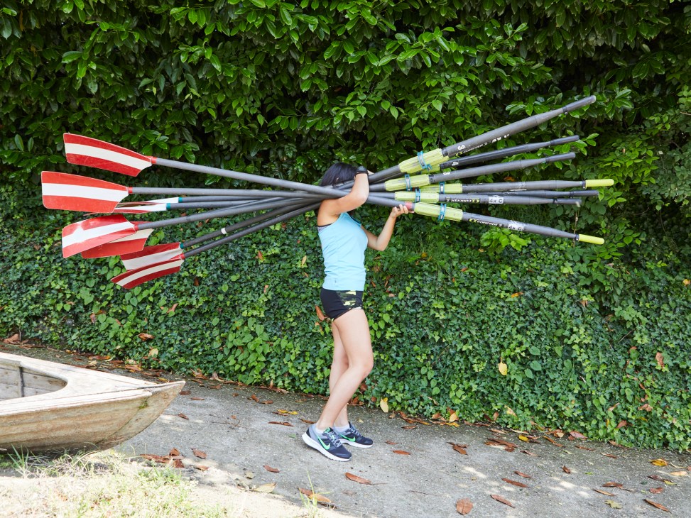 A rower holds multiple oars with red and white striped blades against a leafy green hedge backdrop.