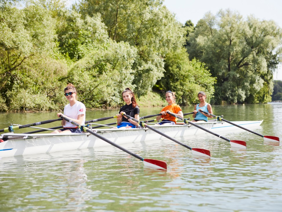 Four female rowers in a boat on the Po River with lush green trees lining the banks.