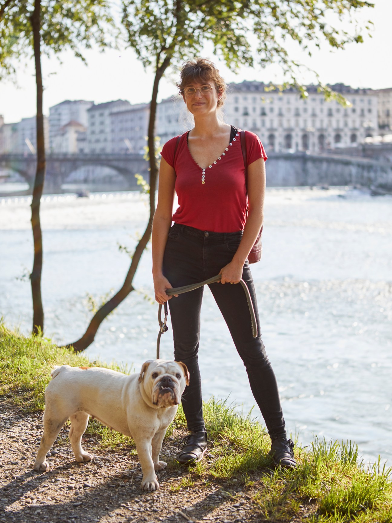 Woman walking a white dog along the Po River in Turin with historic buildings and a bridge visible in the background.