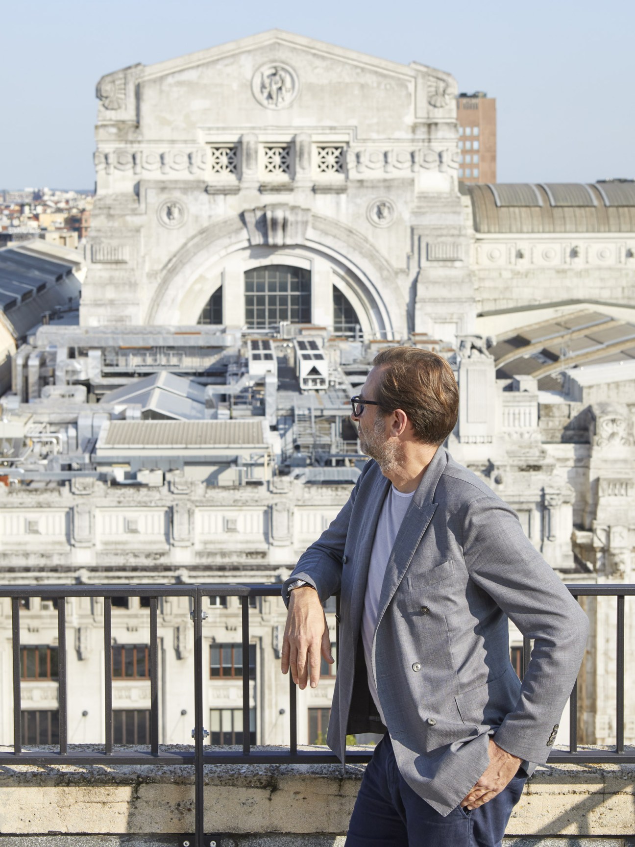 Man in gray blazer and glasses standing on rooftop overlooking classical architecture