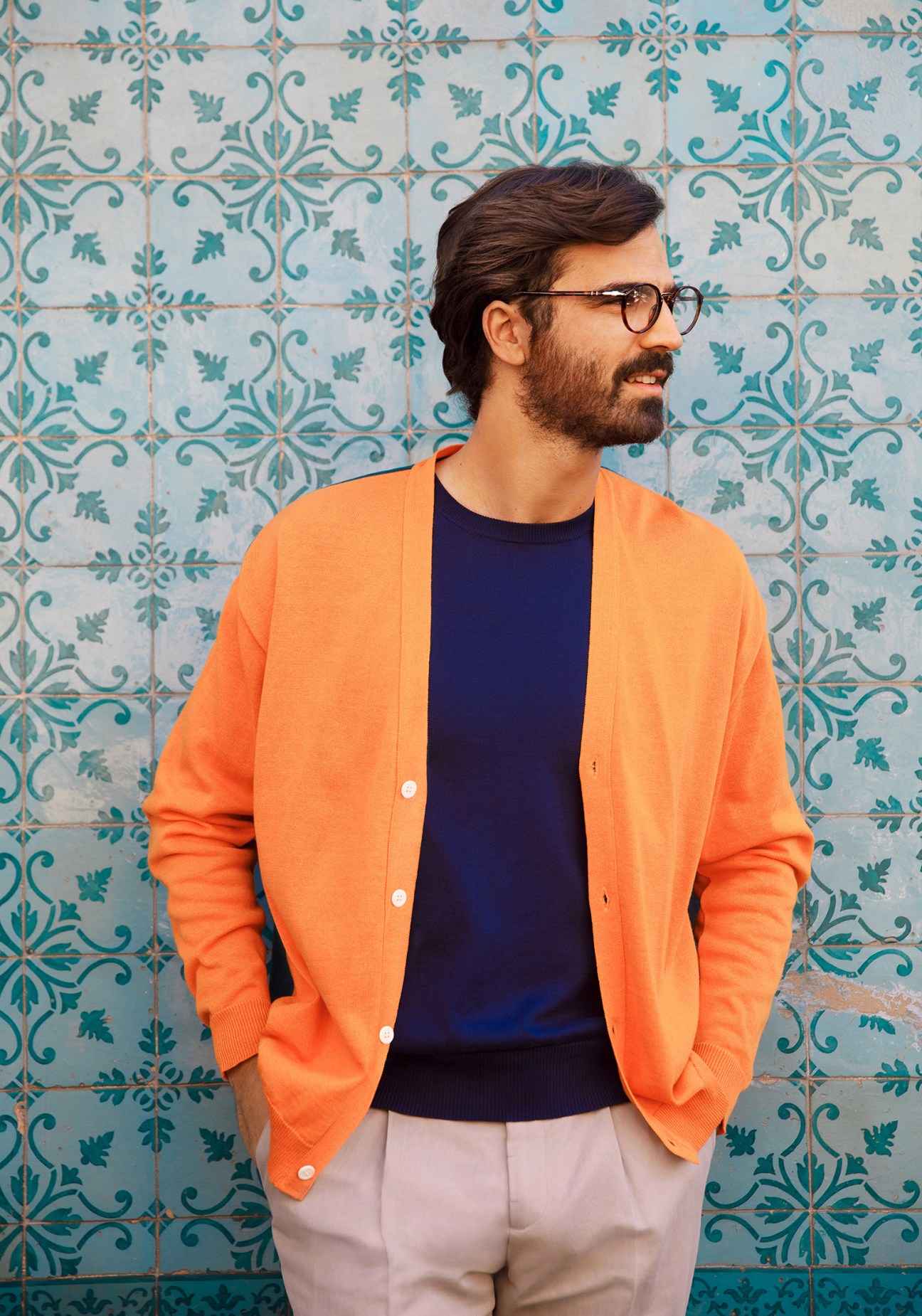Man wearing orange cardigan, navy jumper, and light trousers against turquoise patterned tiles in Lisbon.