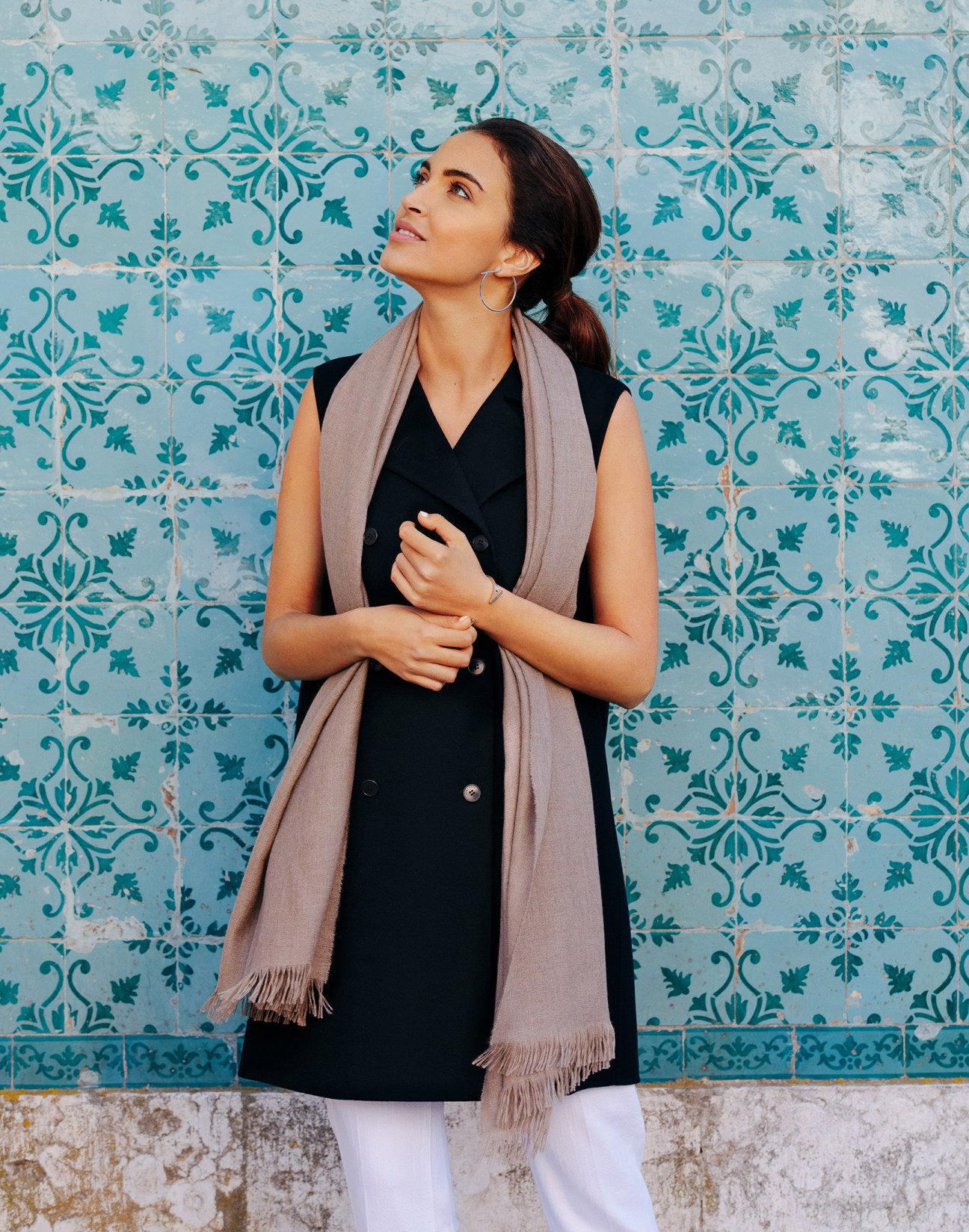 Woman wearing a sleeveless Loro Piana coat and scarf posing against turquoise patterned tiles in Lisbon.
