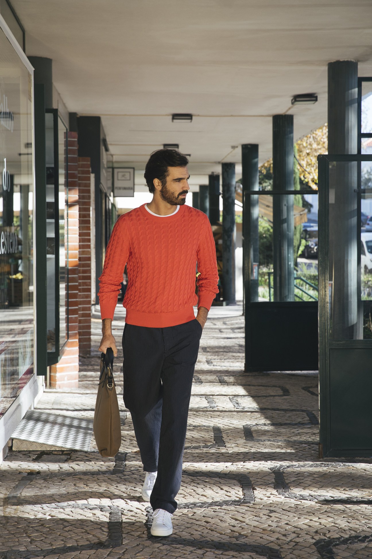 Man in red cable-knit jumper and dark trousers walking through covered shopping arcade carrying tan leather bag.