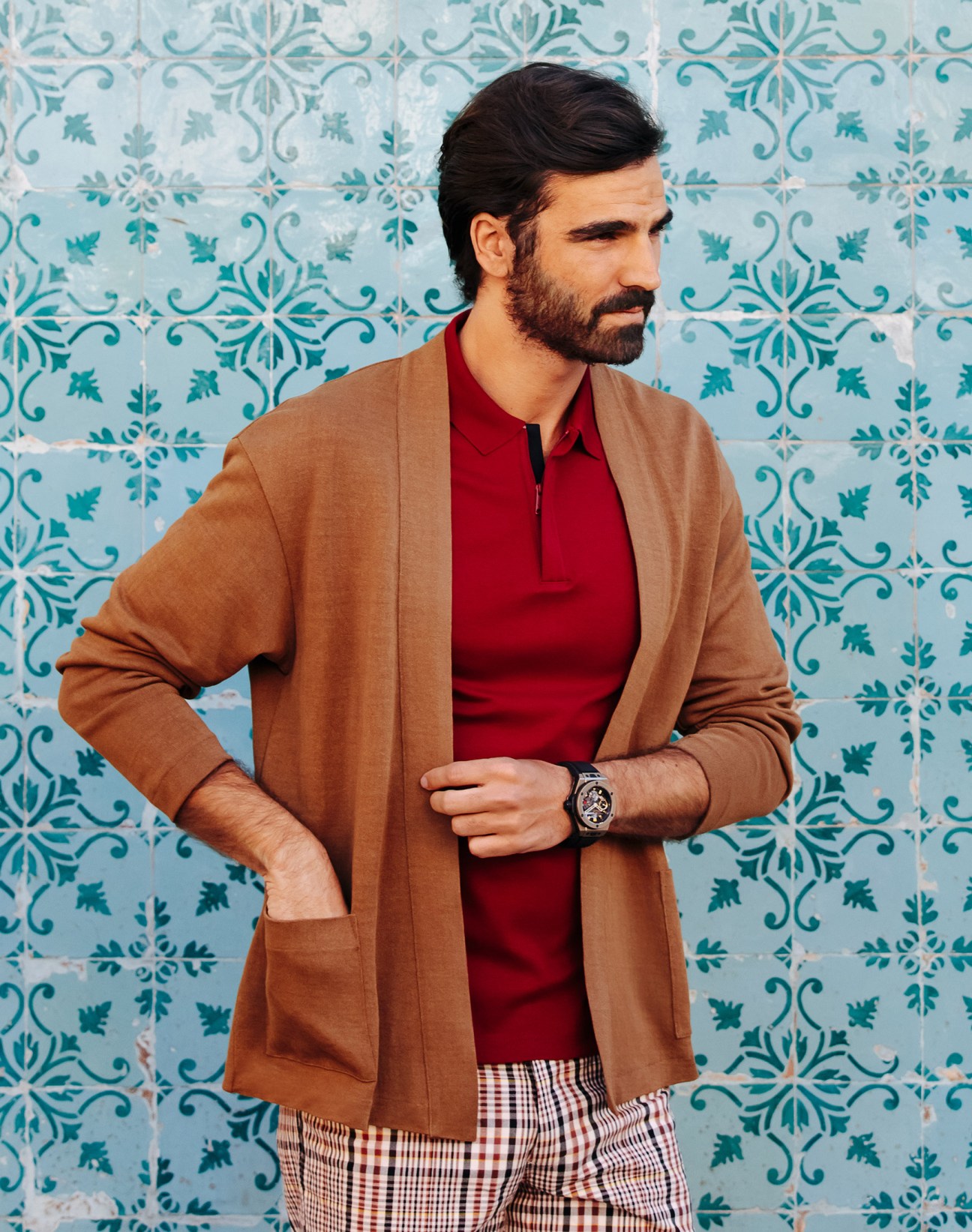 Man wearing camel cardigan and red polo shirt against blue and teal patterned tiles.