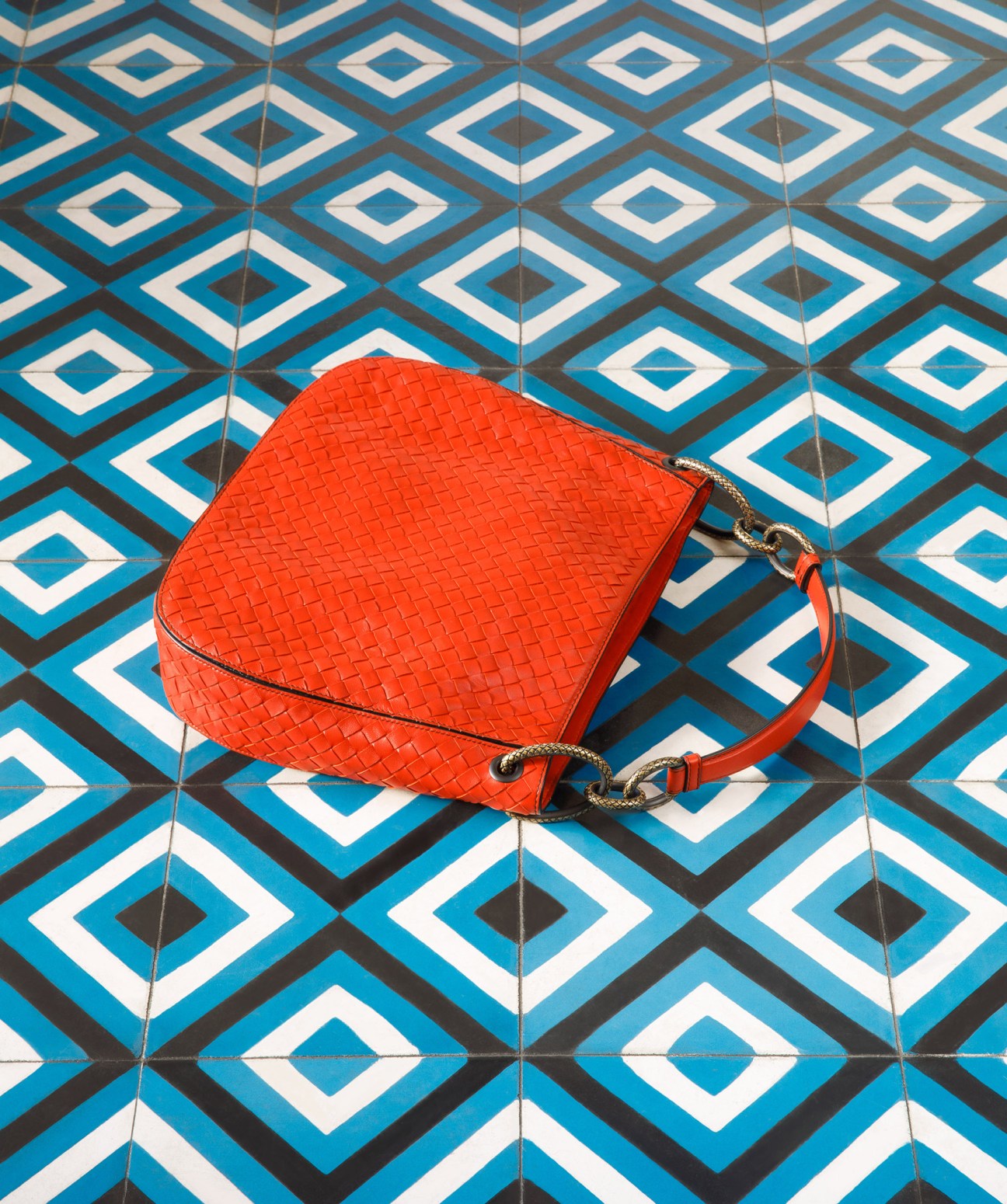 Red woven leather bag by Bottega Veneta on blue and white geometric patterned tiles.