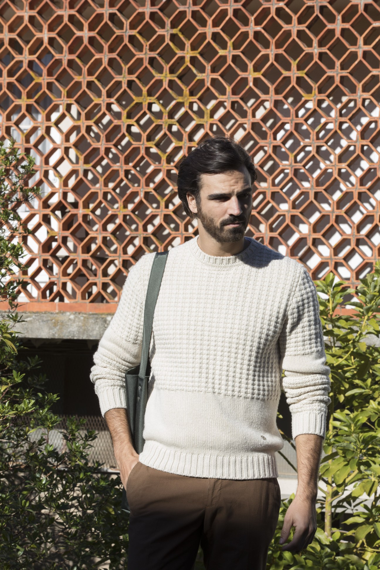Man wearing Louis Vuitton cream jumper with Boglioli trousers and Aeta bag against geometric patterned wall.