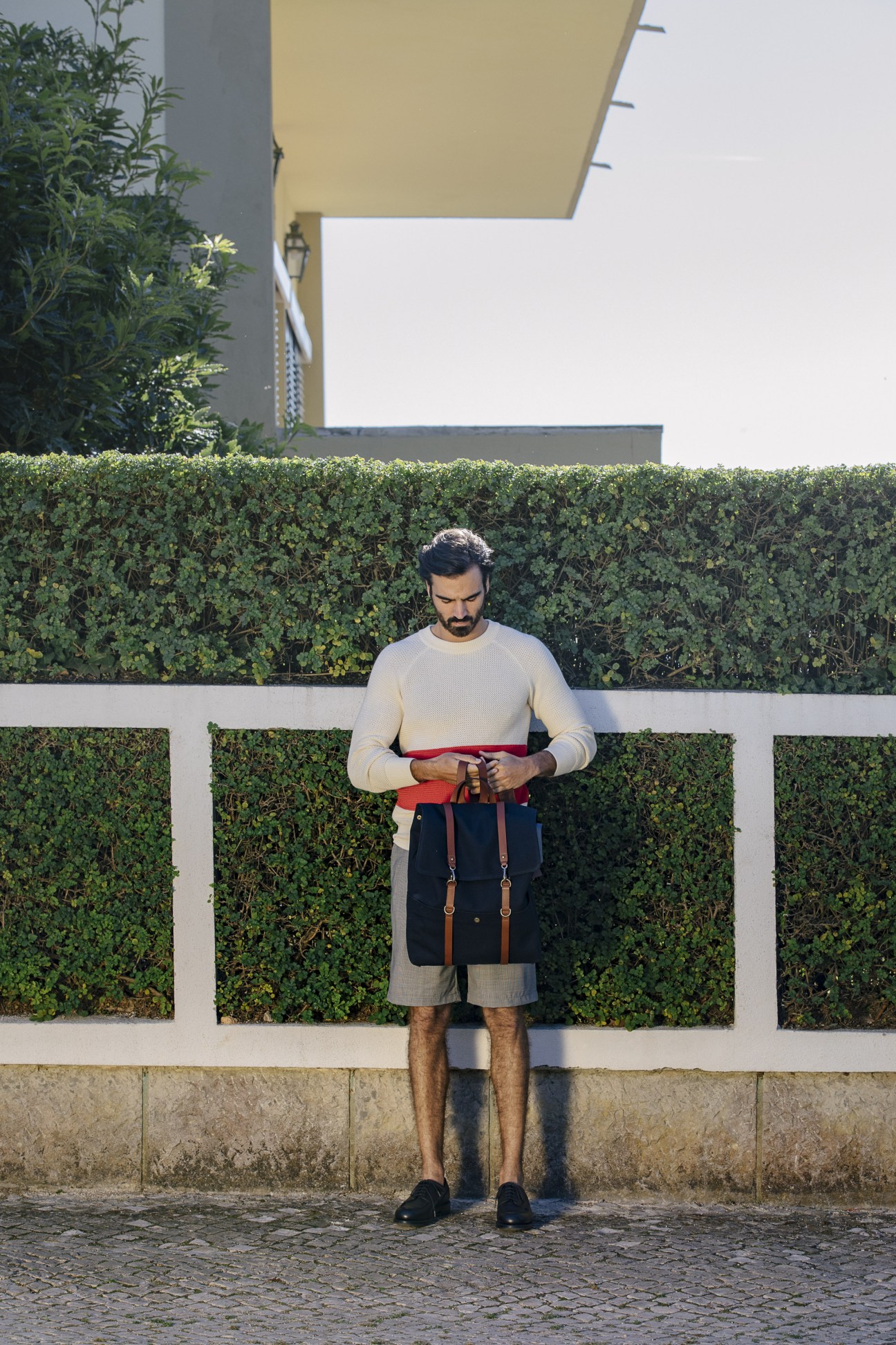 Man wearing La Paz jumper with Universal Products shorts, JM Weston shoes, and Mismo backpack in Lisbon's Restelo district