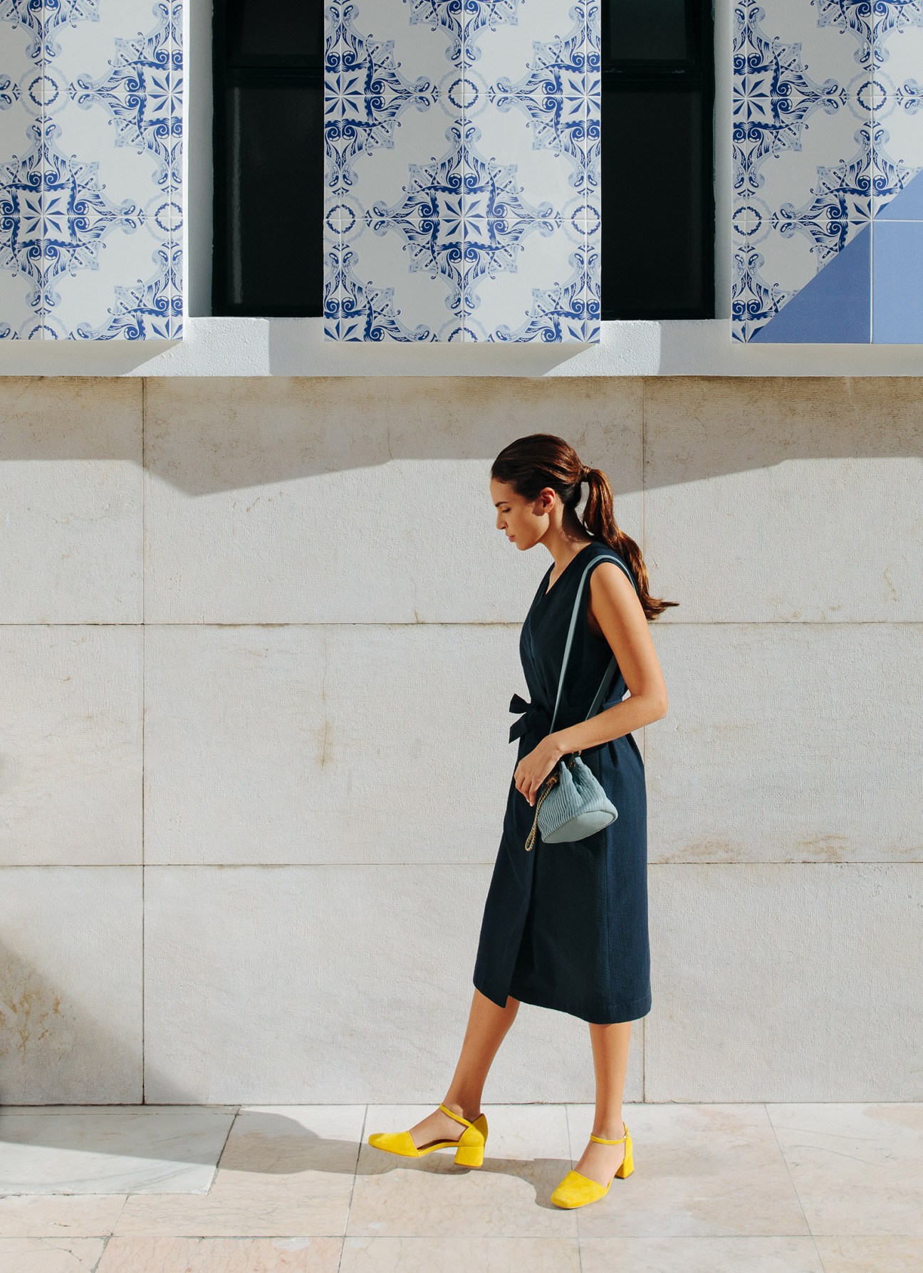 Woman wearing a dark sleeveless dress and yellow block-heel sandals standing against a textured concrete wall with blue and white patterned tiles above.