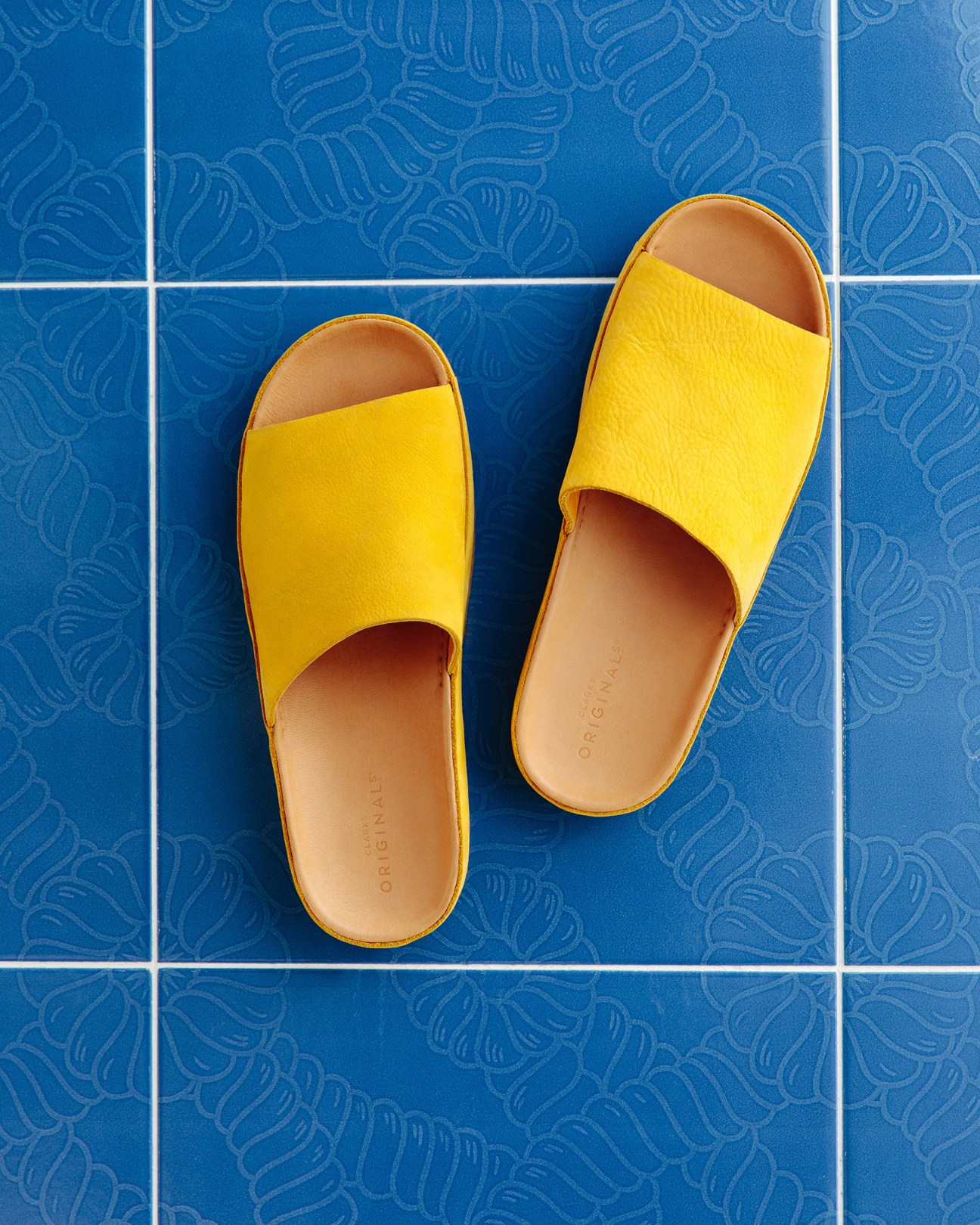 Pair of yellow slides with tan soles on blue patterned tiles by Clarks Originals.