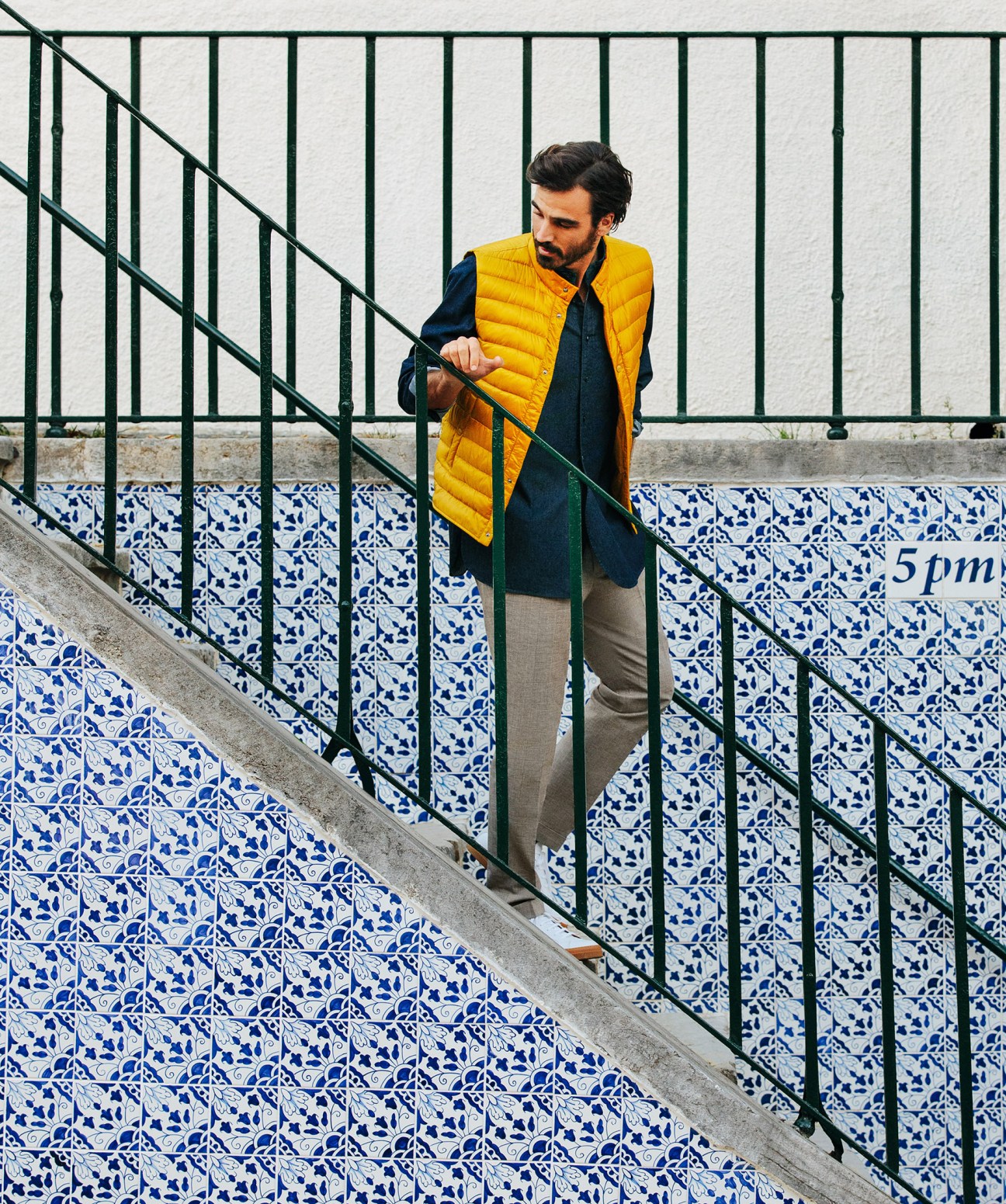 Man wearing yellow gilet by Herno and navy shirt on Lisbon stairs with blue and white azulejo tiles.