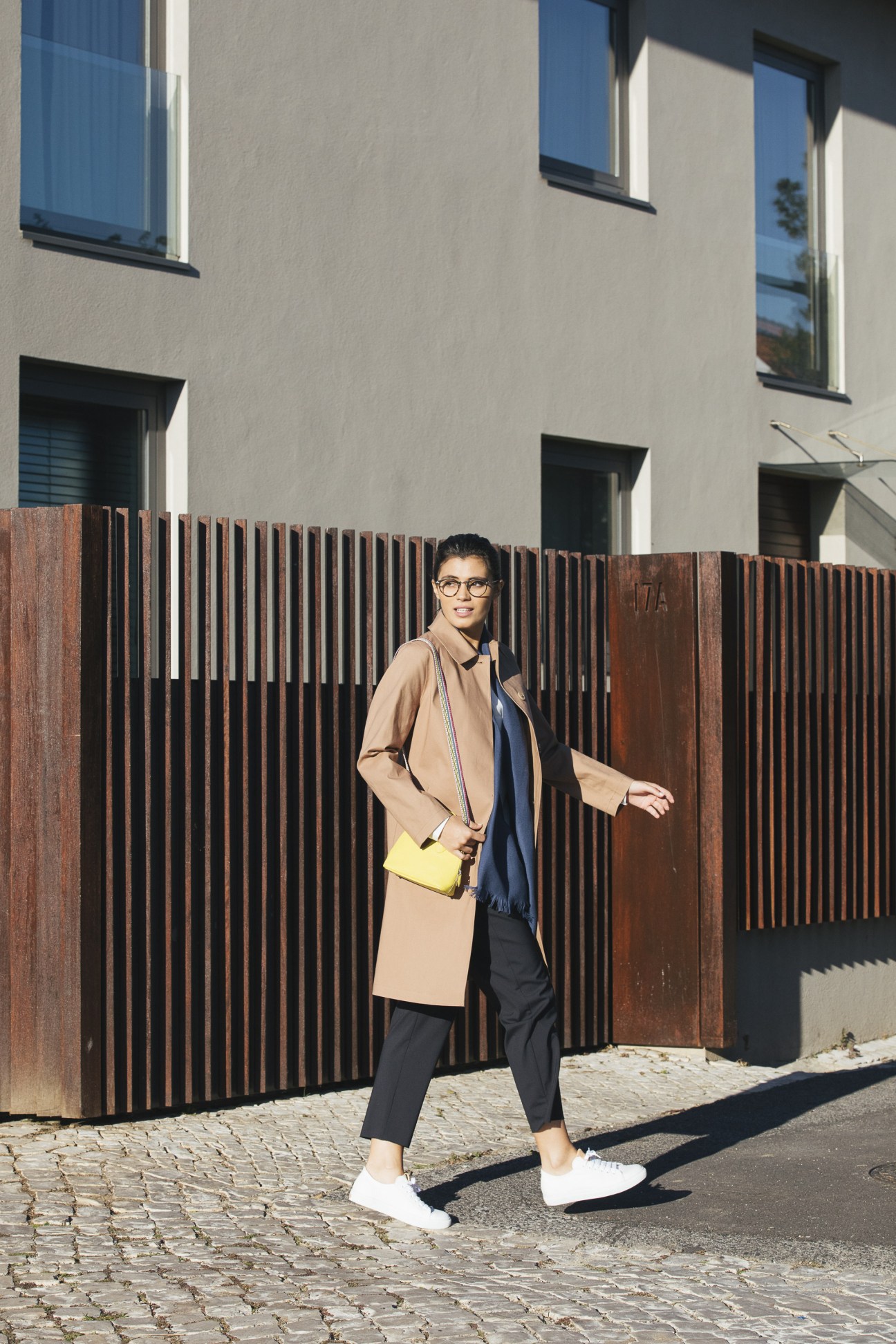 Model wearing Mackintosh coat with Sunspel blouse and Barena trousers, accessorized with Closed trainers, Oliver Peoples glasses, Begg & Co stole, and Hermès bag