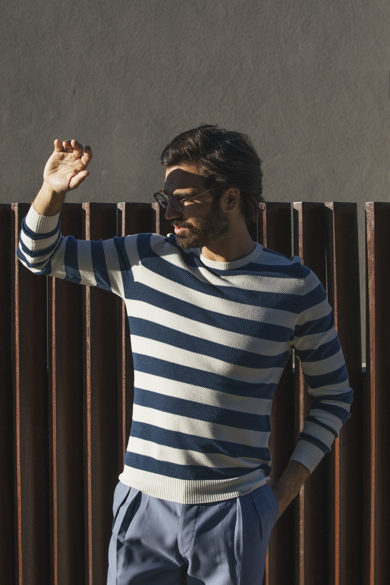 Model wearing Brooksfield striped jumper with Orazio Luciano trousers and Prada glasses against a corrugated metal wall.