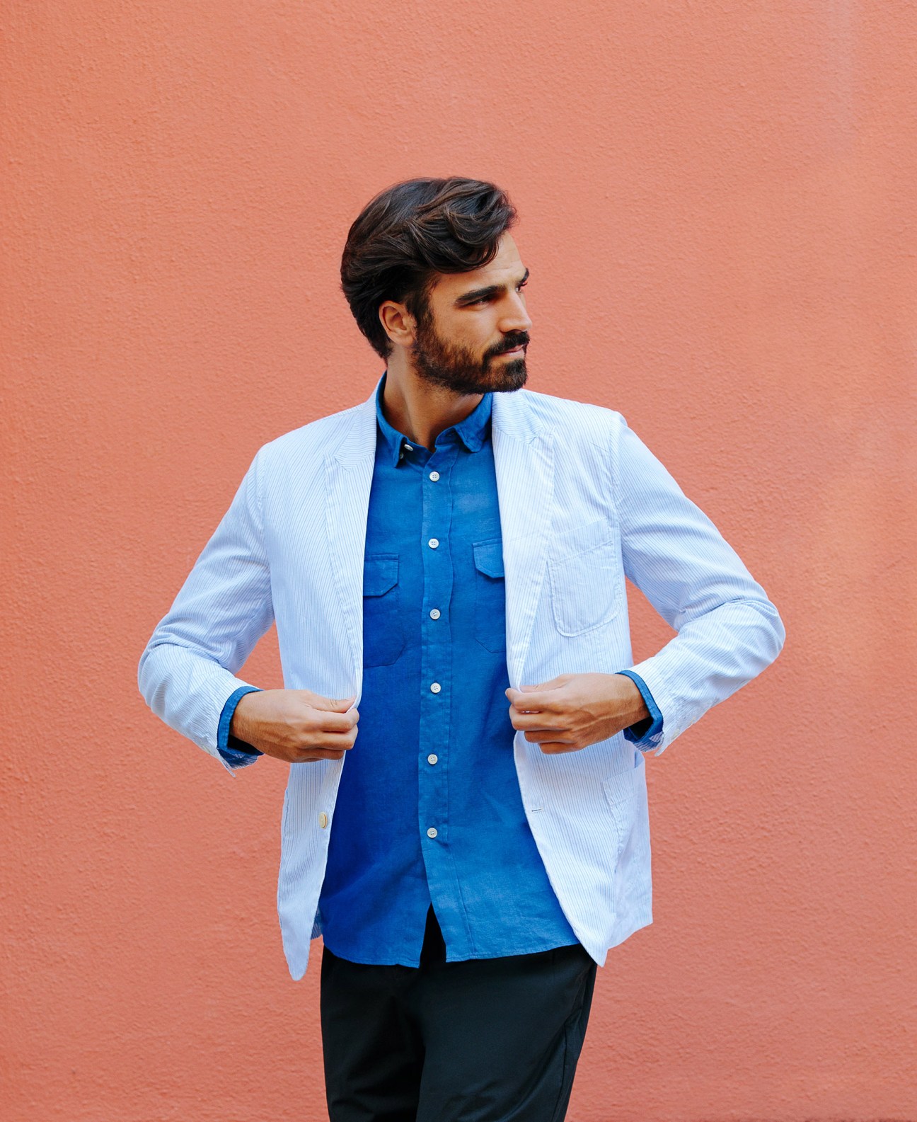 Man wearing light blue jacket by Roy Roger's, blue shirt by La Paz, and dark trousers by A Kind of Guise against coral background.