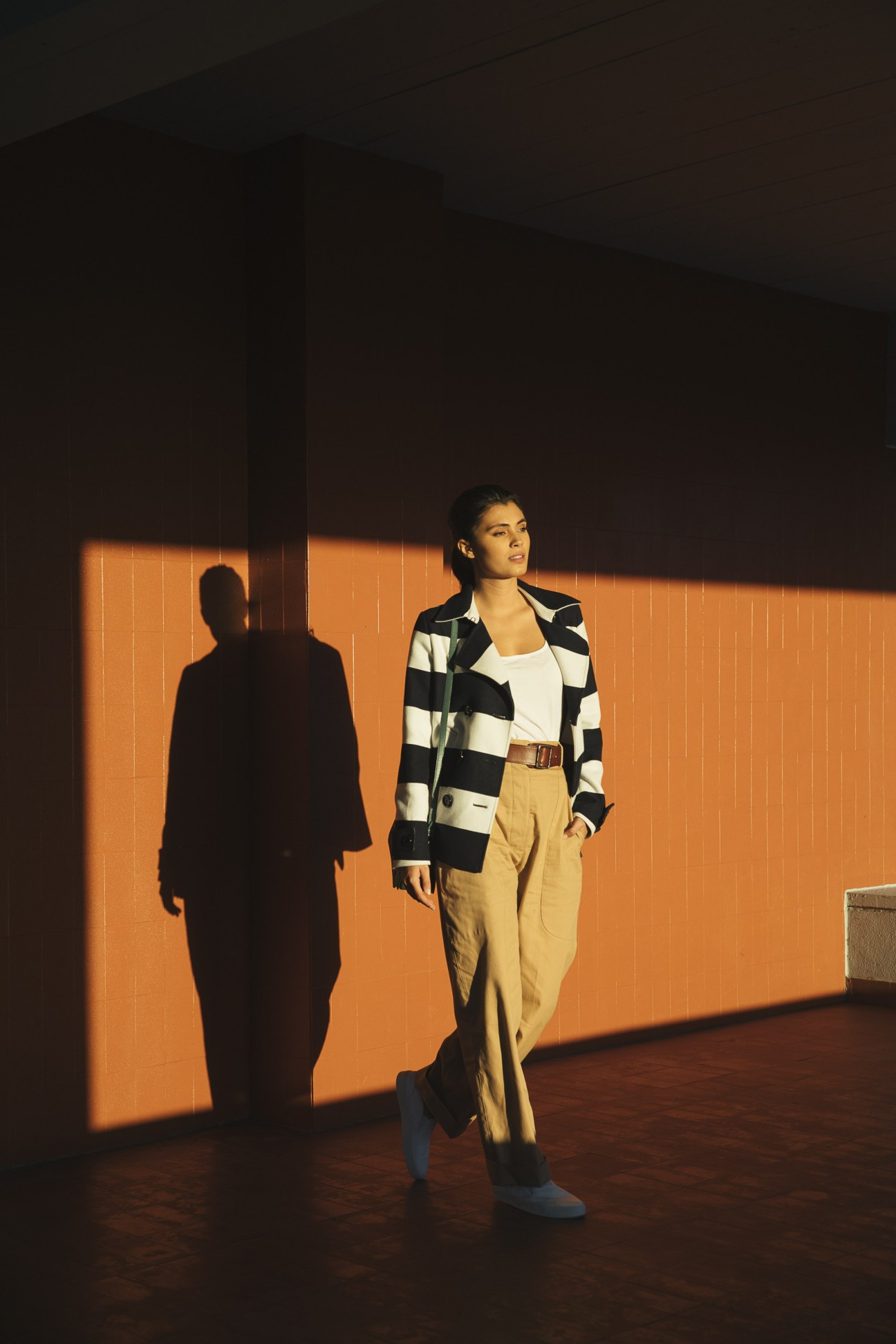 Model in black and white checked Herno coat with beige trousers against orange-lit wall in Lisbon.