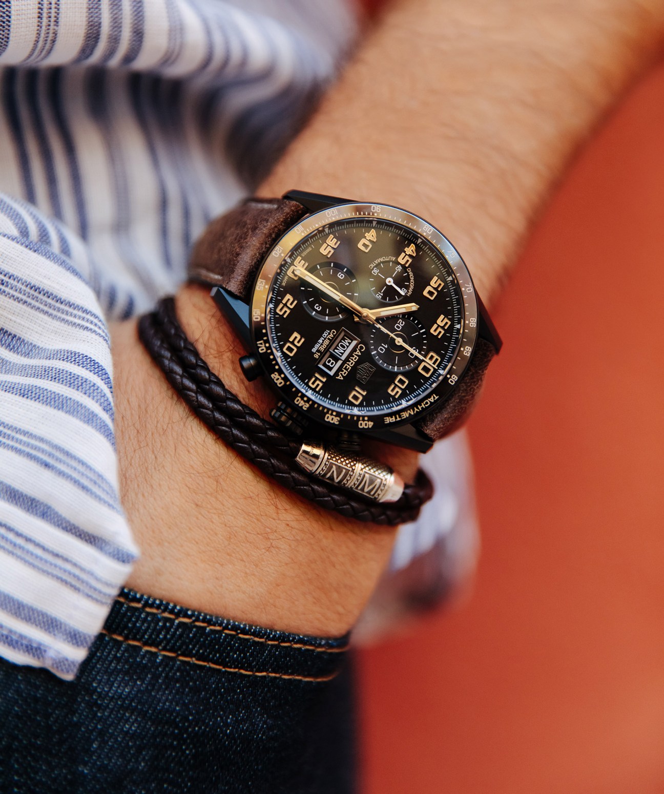 Man's wrist wearing a Tag Heuer chronograph watch with brown leather strap and a Tateossian bracelet, against an orange background.