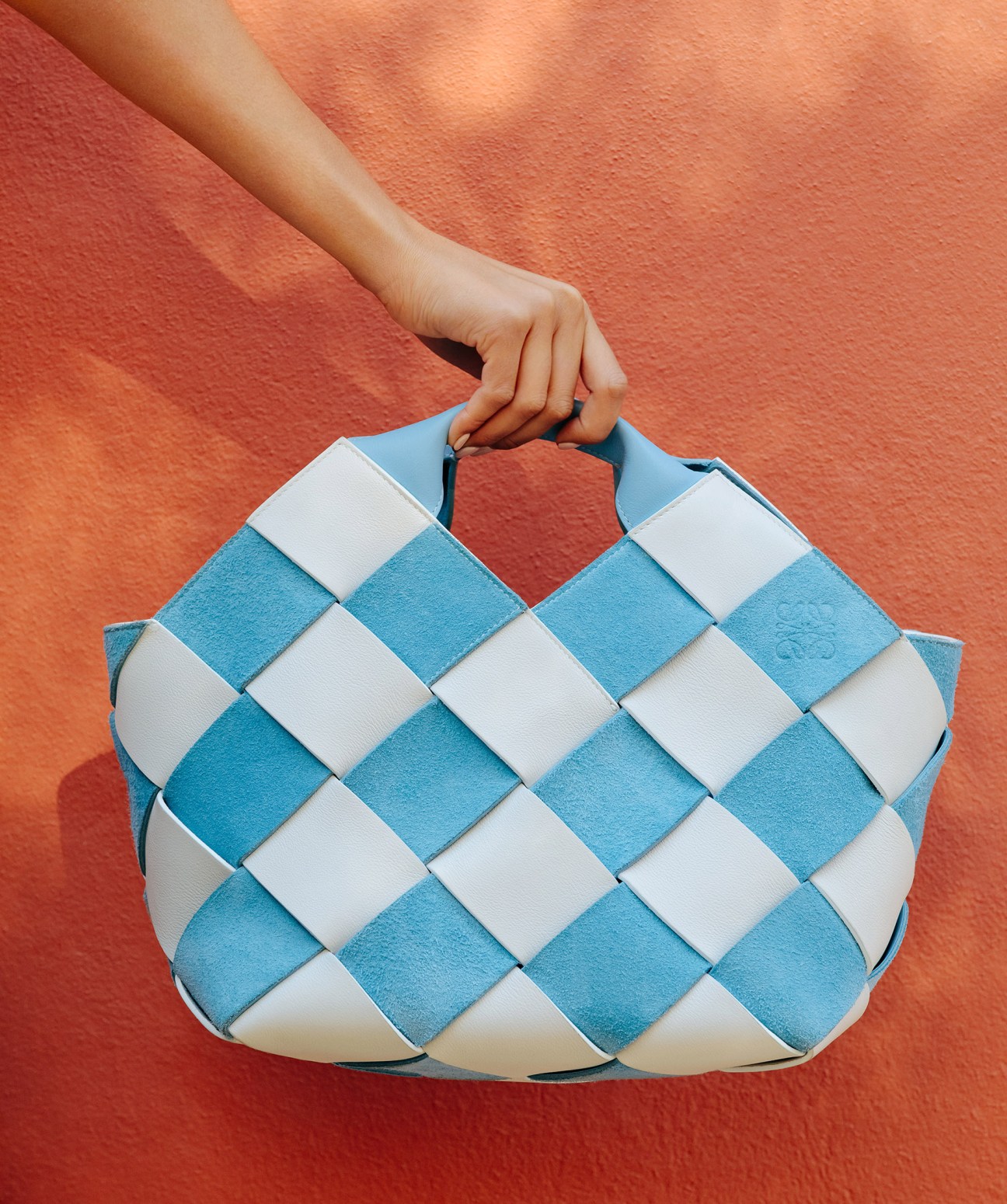 A person holds a blue and white woven leather bag by Loewe against a terracotta-colored wall.