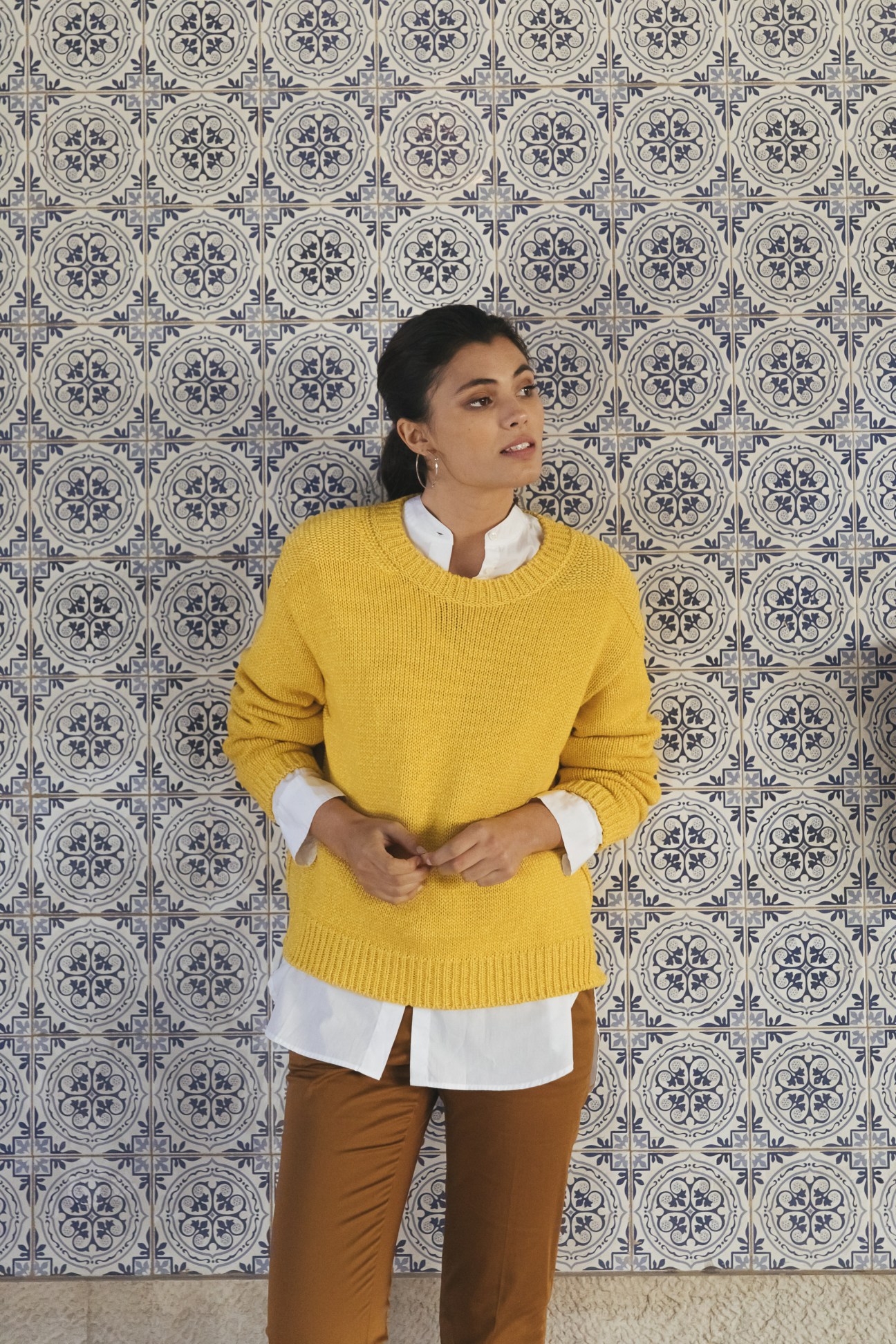 Model in yellow Closed jumper with white Arket shirt and brown PT Pantaloni Torino trousers against blue patterned tiles.