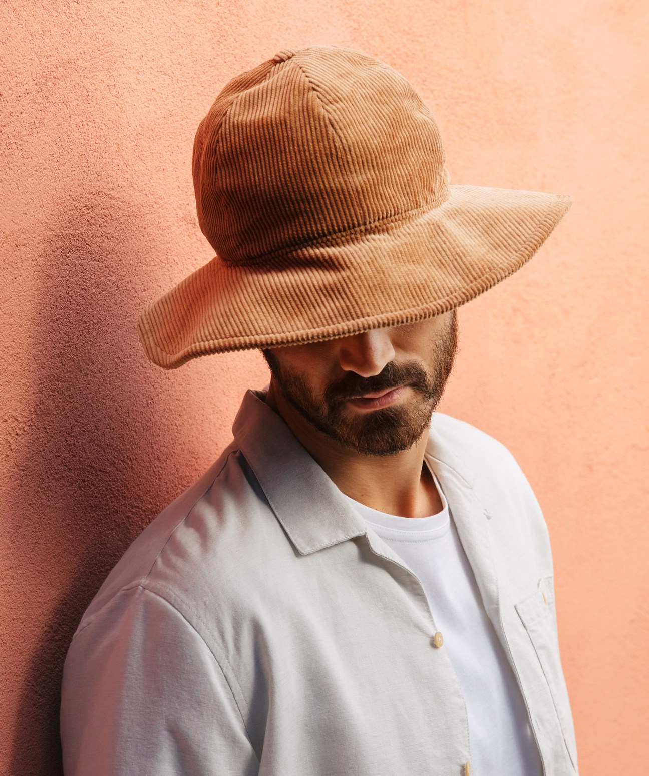 Man wearing beige hat by A Kind of Guise and white t-shirt against pink textured wall in Lisbon.