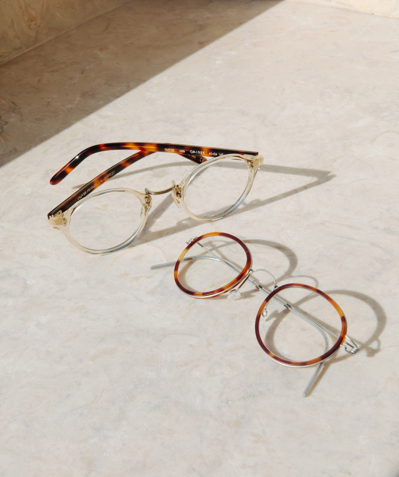 Three eyeglass frames displayed on a white marble surface: tortoiseshell and clear frames by Oliver Peoples and Lindberg.